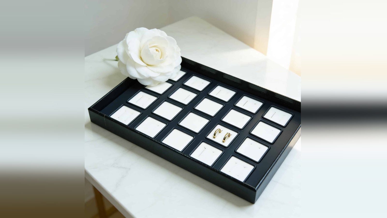 Bandeja forrada con compartimentos y piezas de joyería, sobre mármol blanco con una rosa